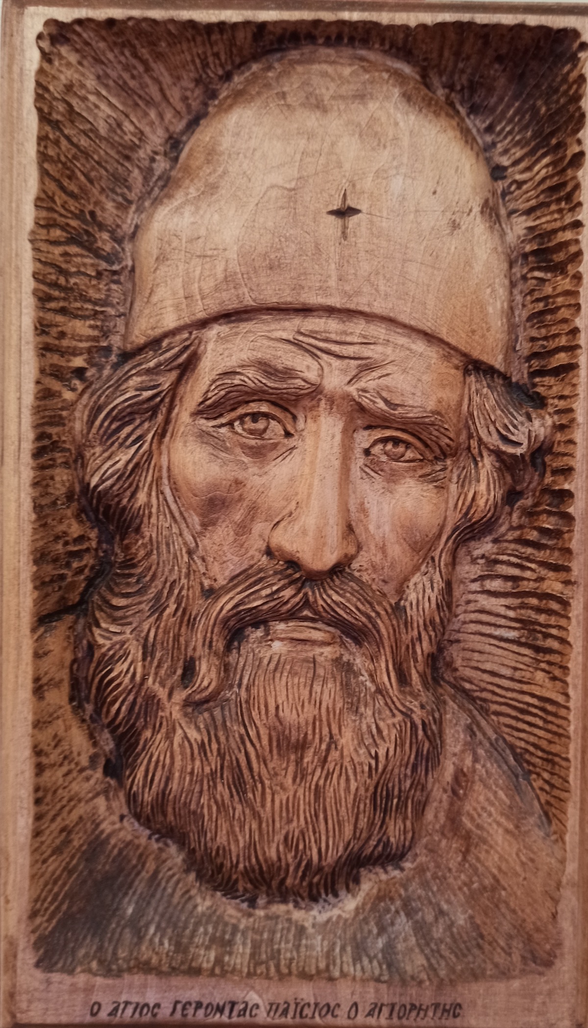 Wood-carved portrait of St. Paisios the Athonite, showing his distinctive monastic cap and gentle, weathered face, with Greek inscription
